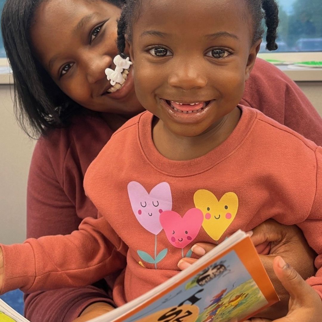 Young child smiling while sitting with an adult and holding a children’s book during a shared reading moment.